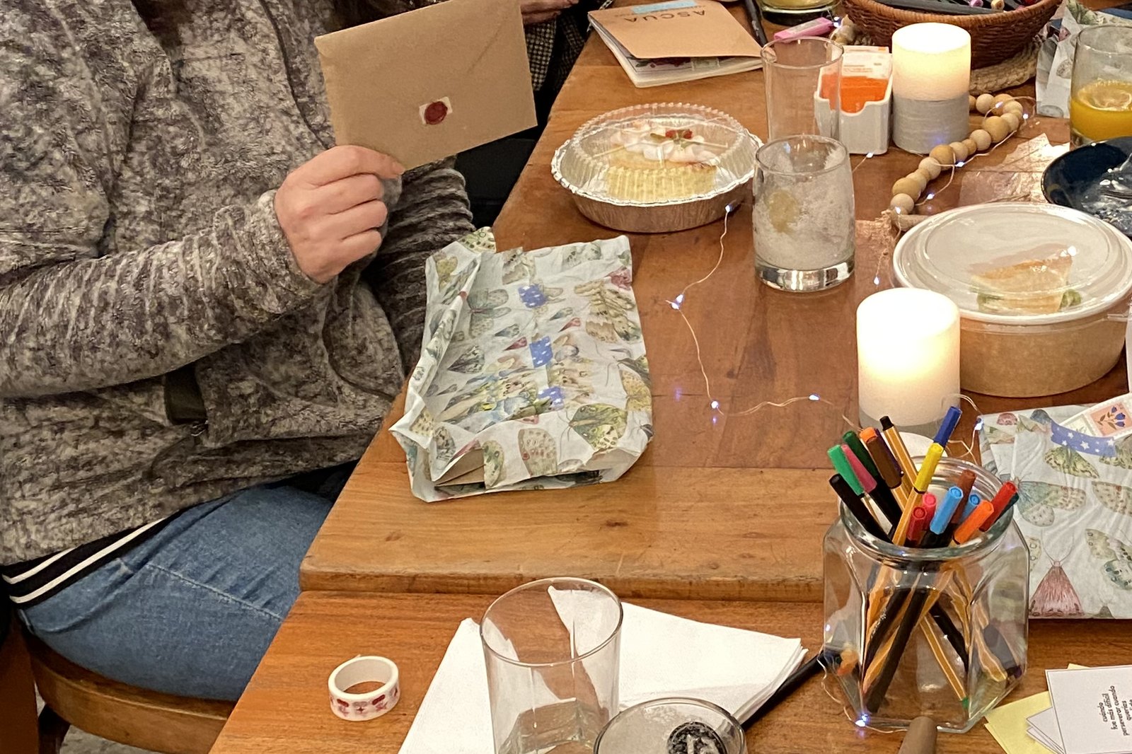 La mesa larga del primer encuentro, con manteles de mariposas, velas, un frasco con lápices de colores y una carta cerrada con lacre en la mano de una participante.